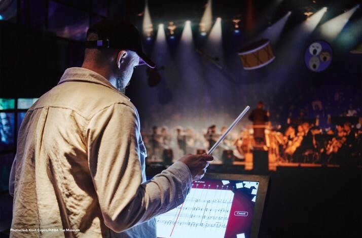 A man conducting and an orchestra on the background. © Knut Capra / Abba The Museum