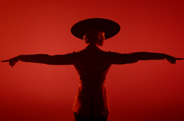 Performer in wide-brimmed hat, arms outstretched, silhouetted against red background.