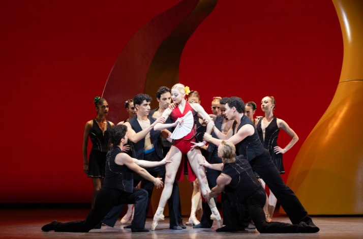 Group of dancers surround central figure in red, dynamic stage interaction.