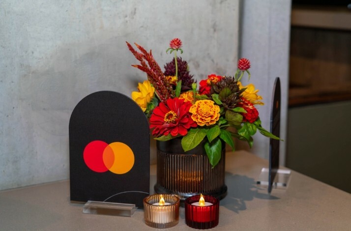 Stand with Mastercard logo next to flowers and candles in Mastercard colors on a table at the Moody Center.